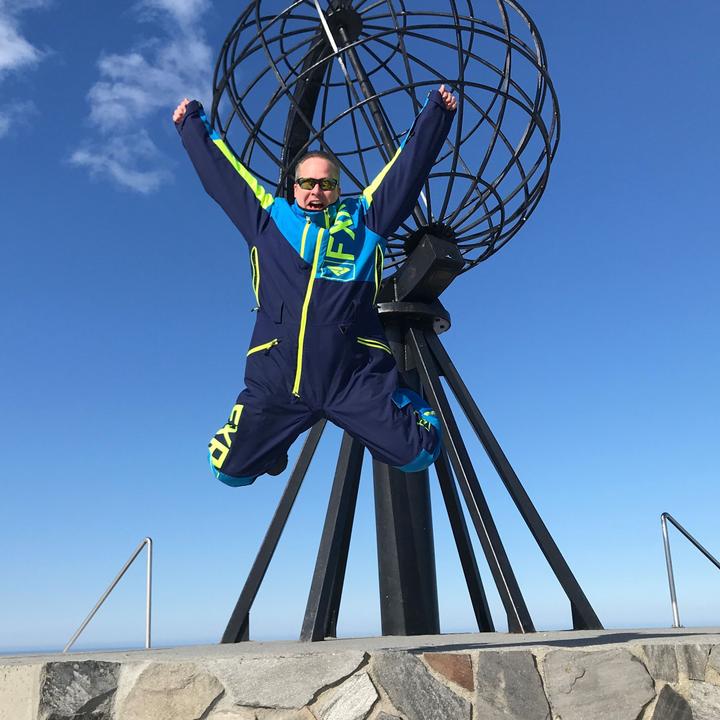 Ein Besucher springt mit erhobenen Armen vor dem ikonischen Nordkap-Globusdenkmal in den klaren blauen arktischen Himmel und feiert an einem der nördlichsten Wahrzeichen Europas.