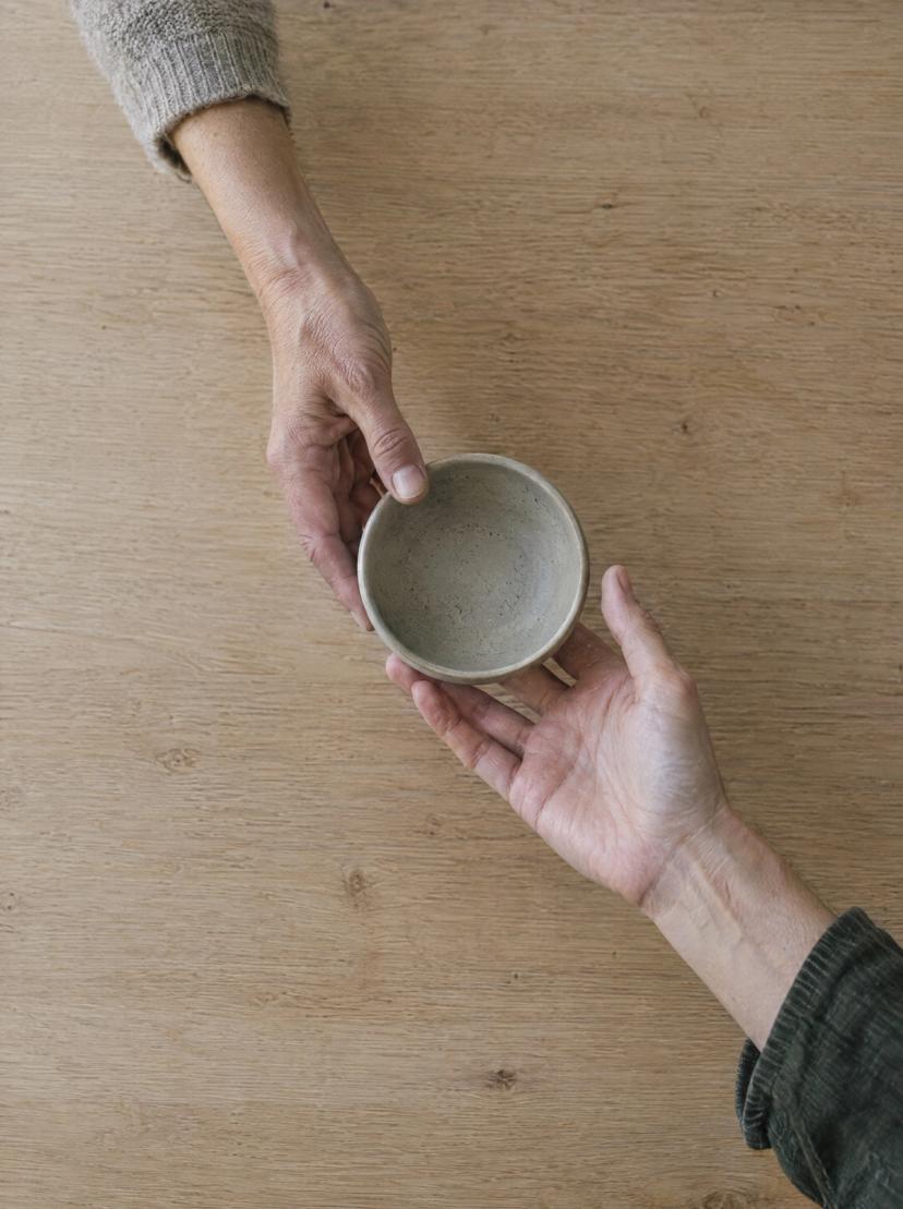 One hand passing a small ceramic bowl to another hand across an oak table, seen from above
