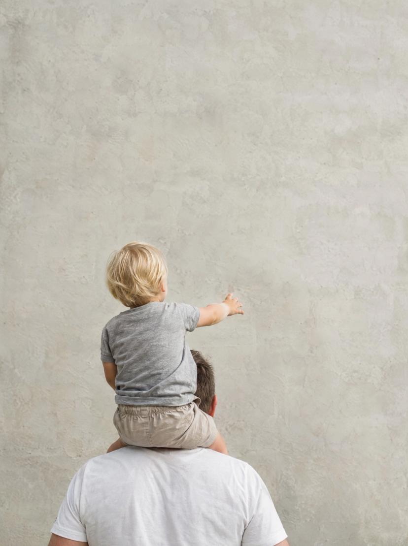 Un adulto lleva a un niño a hombros junto a una pared de hormigón clara, vistos de espaldas