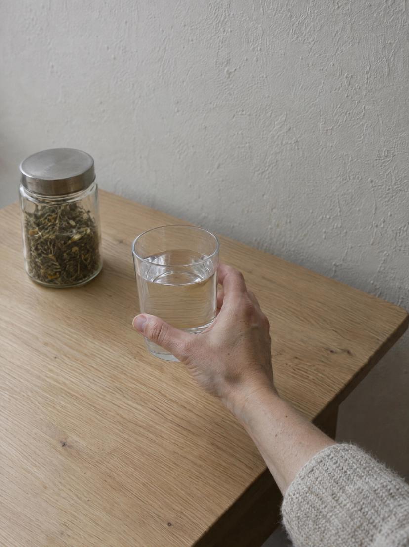 Una mano levanta un vaso de agua de una mesa de roble, con un tarro de infusión apartado a un lado