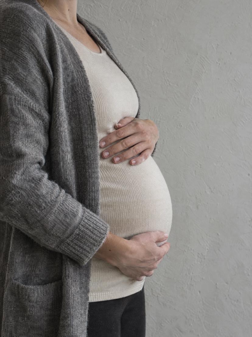 A pregnant woman standing with her hands on her belly by a pale wall, seen from the side