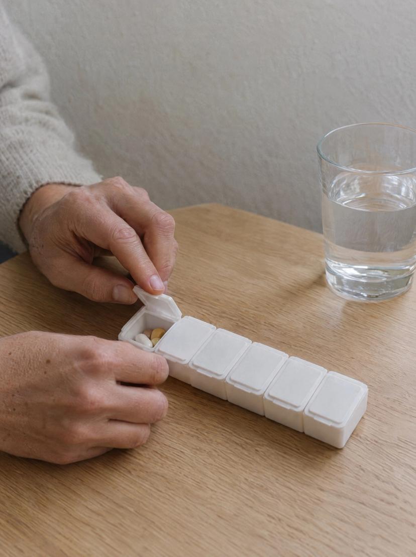 Hands opening one compartment of a white pill organiser on an oak table, with a glass of water beside it