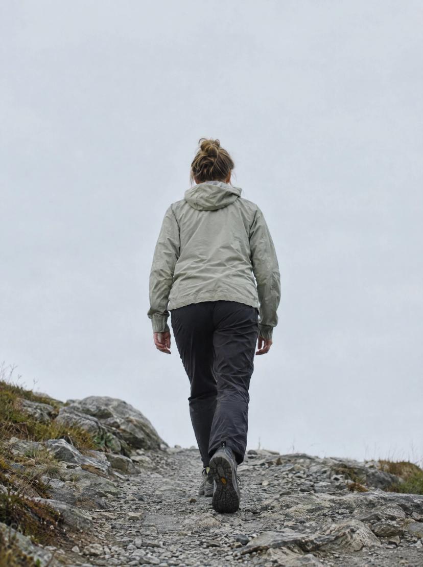 A person in a light jacket walking up a rocky path, seen from behind against a pale sky