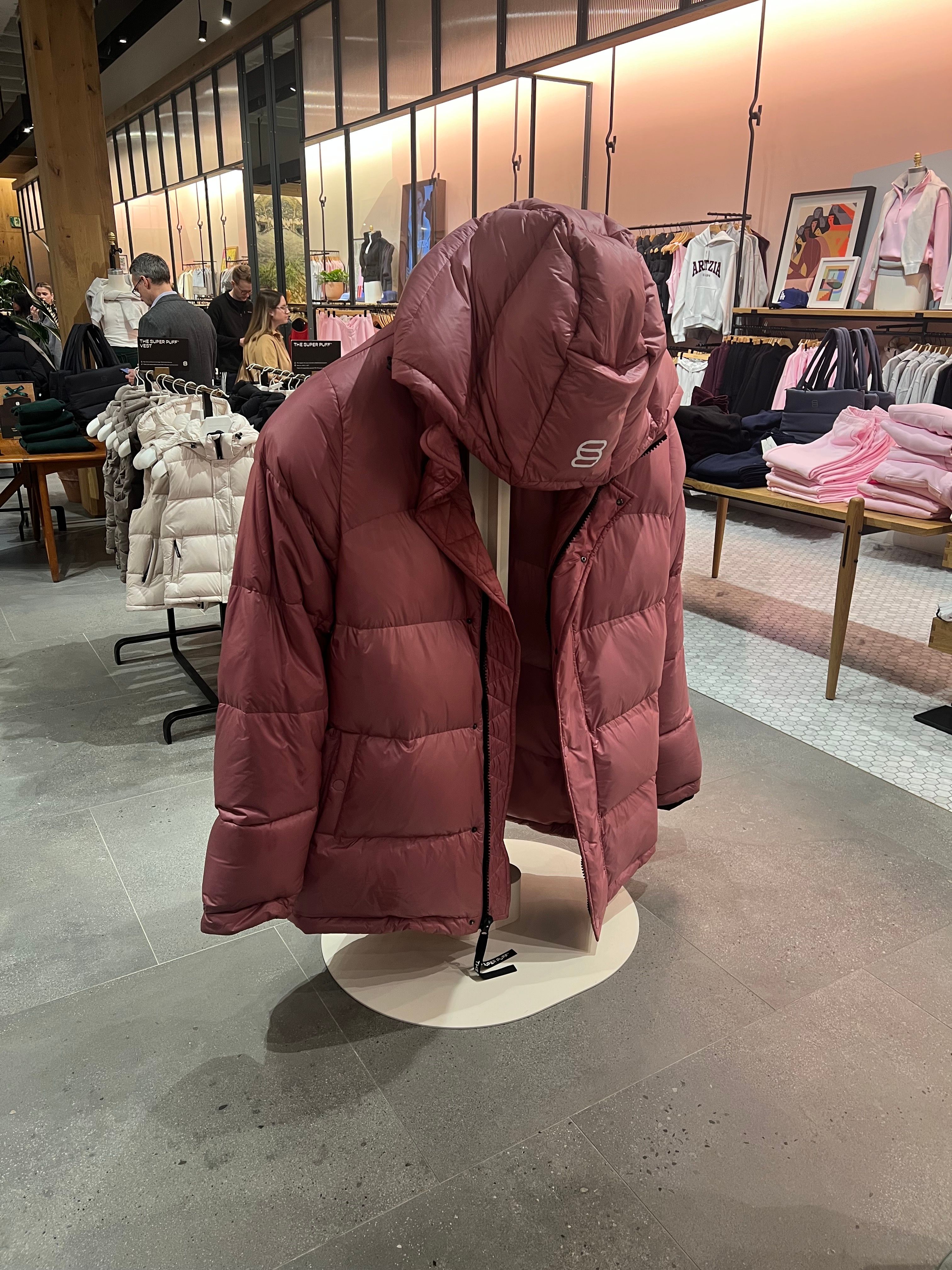 A mauve puffer jacket displayed on a rotating stand in a clothing store, with tables of folded garments, hanging clothes, and shoppers in the background.