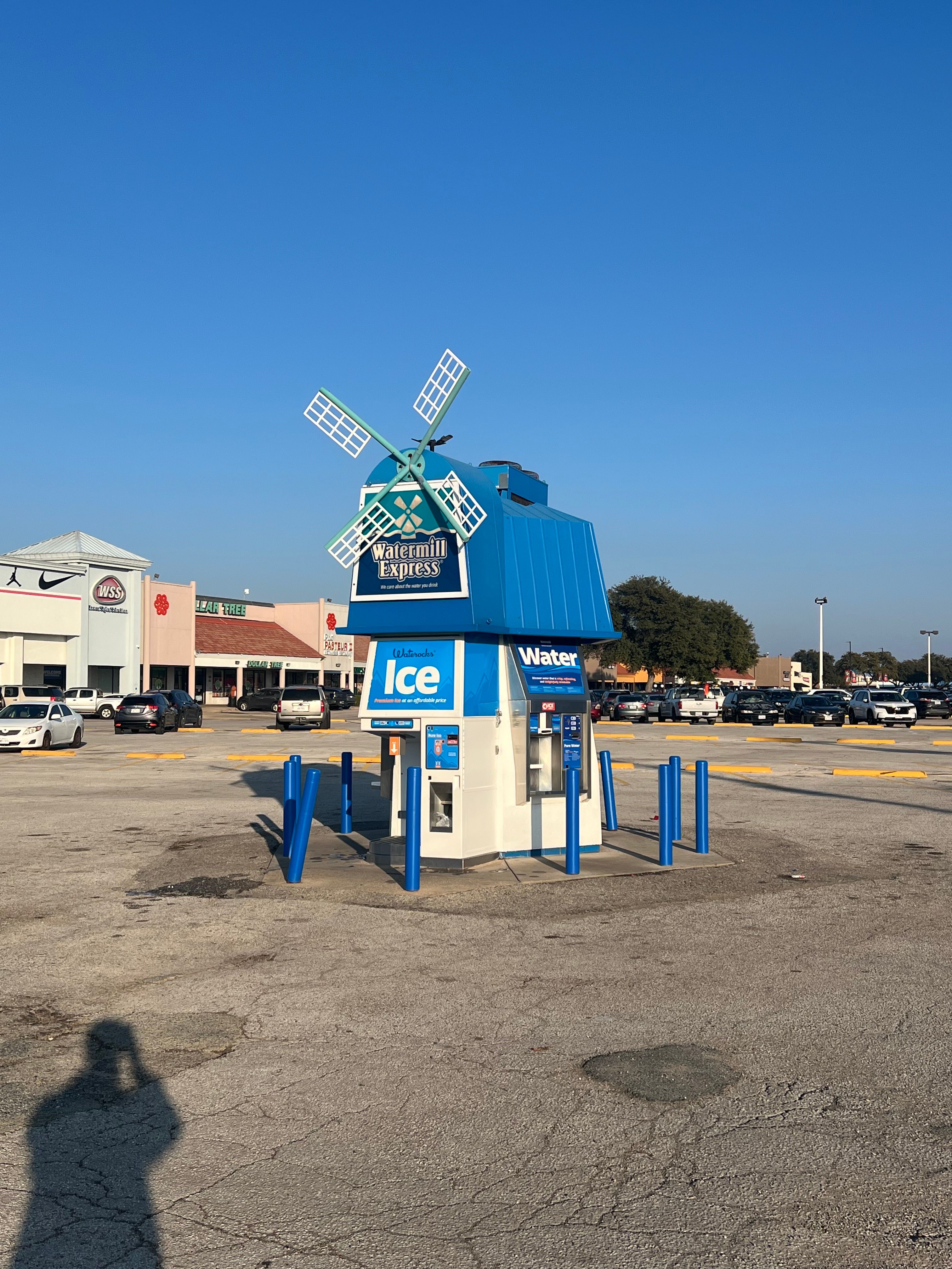 A blue windmill-shaped Watermill Express ice and water vending kiosk surrounded by blue bollards in an empty parking lot with stores in the background under a clear sky.