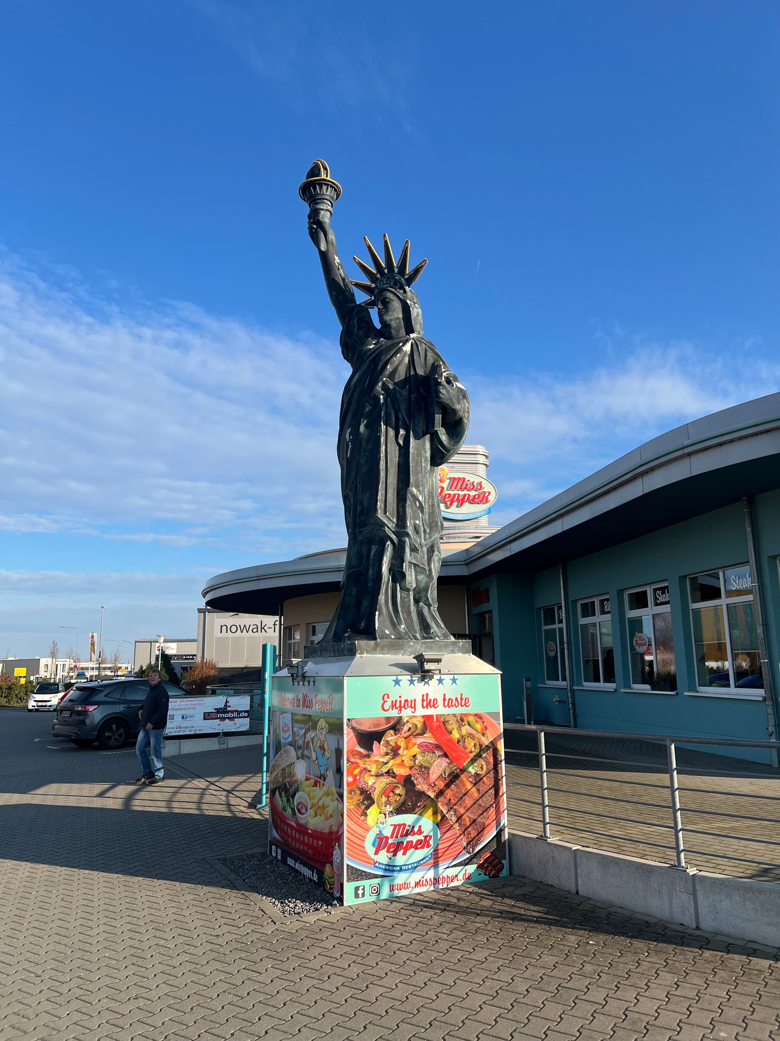 A dark replica of the Statue of Liberty stands on a pedestal wrapped in Miss Pepper restaurant advertising in front of a teal-colored building under a blue sky.