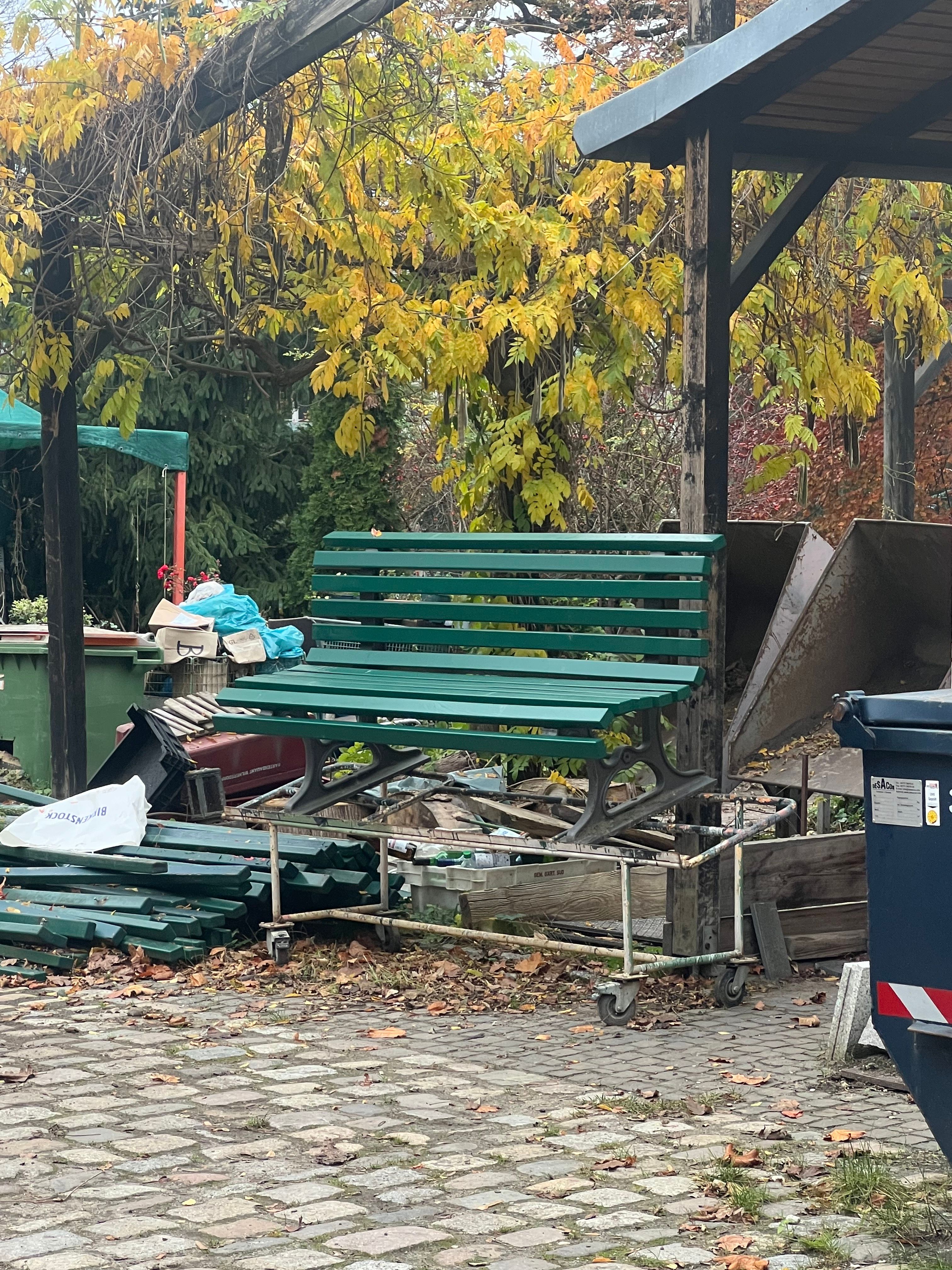 A green wooden bench stacked on a metal cart in a cluttered outdoor storage area with trash bins, wooden planks, and autumn foliage in Berlin.