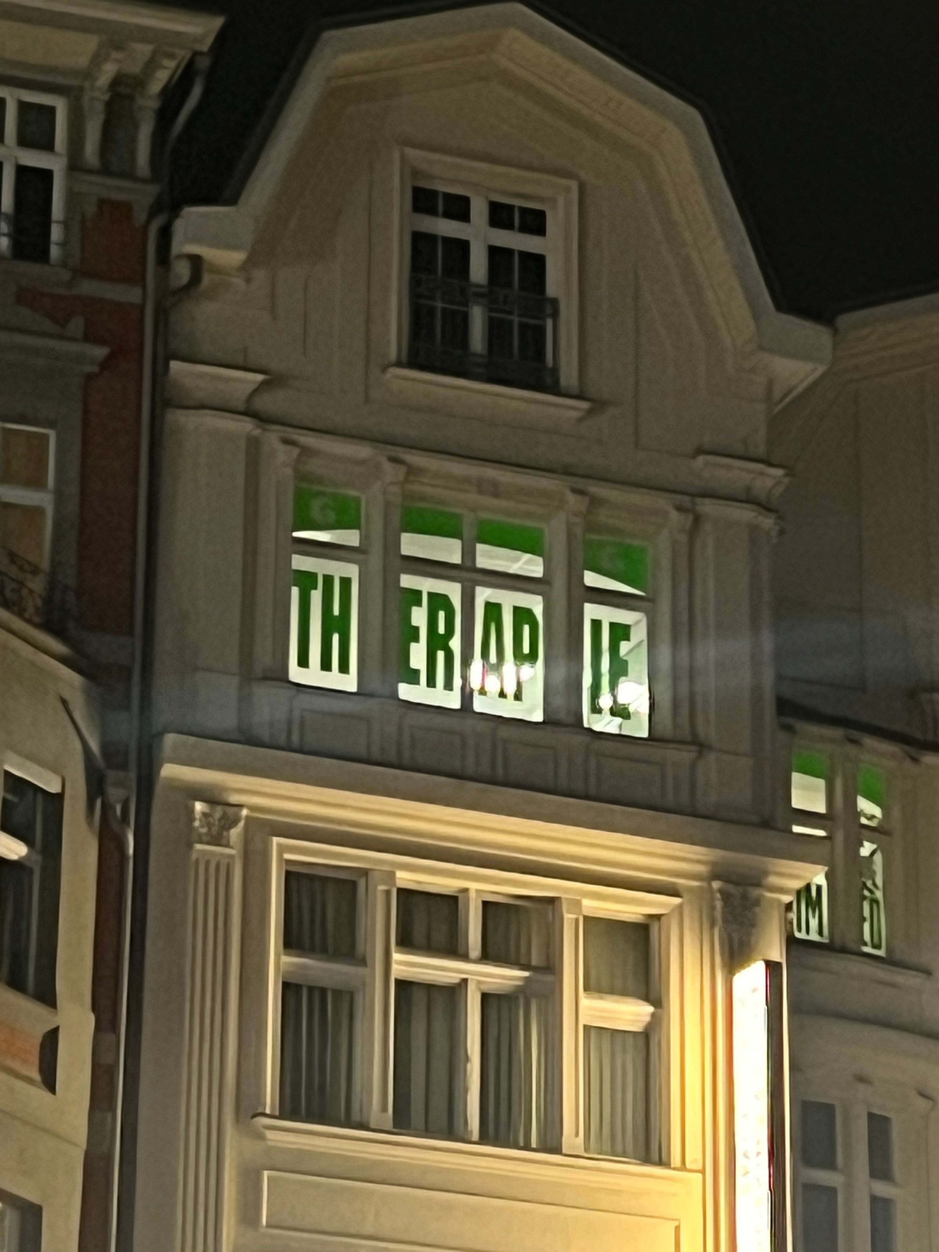 A pale multi-story building facade at night with the word THERAPIE spelled out in green letters across illuminated upper-floor windows.