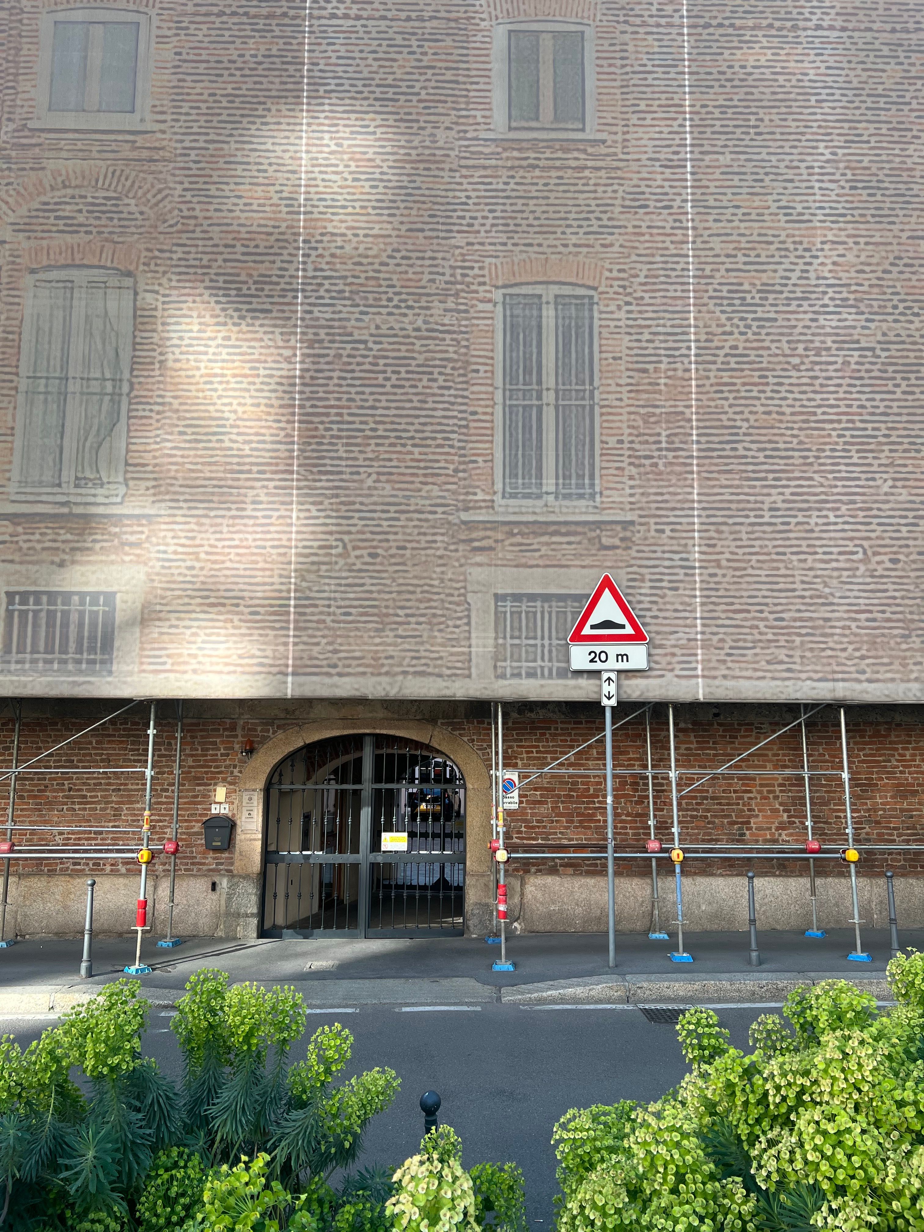 A large brick building with a printed facade covering is fronted by metal scaffolding, an arched gated doorway, and a triangular road bump warning sign reading 20 m, with green plants in the foreground.