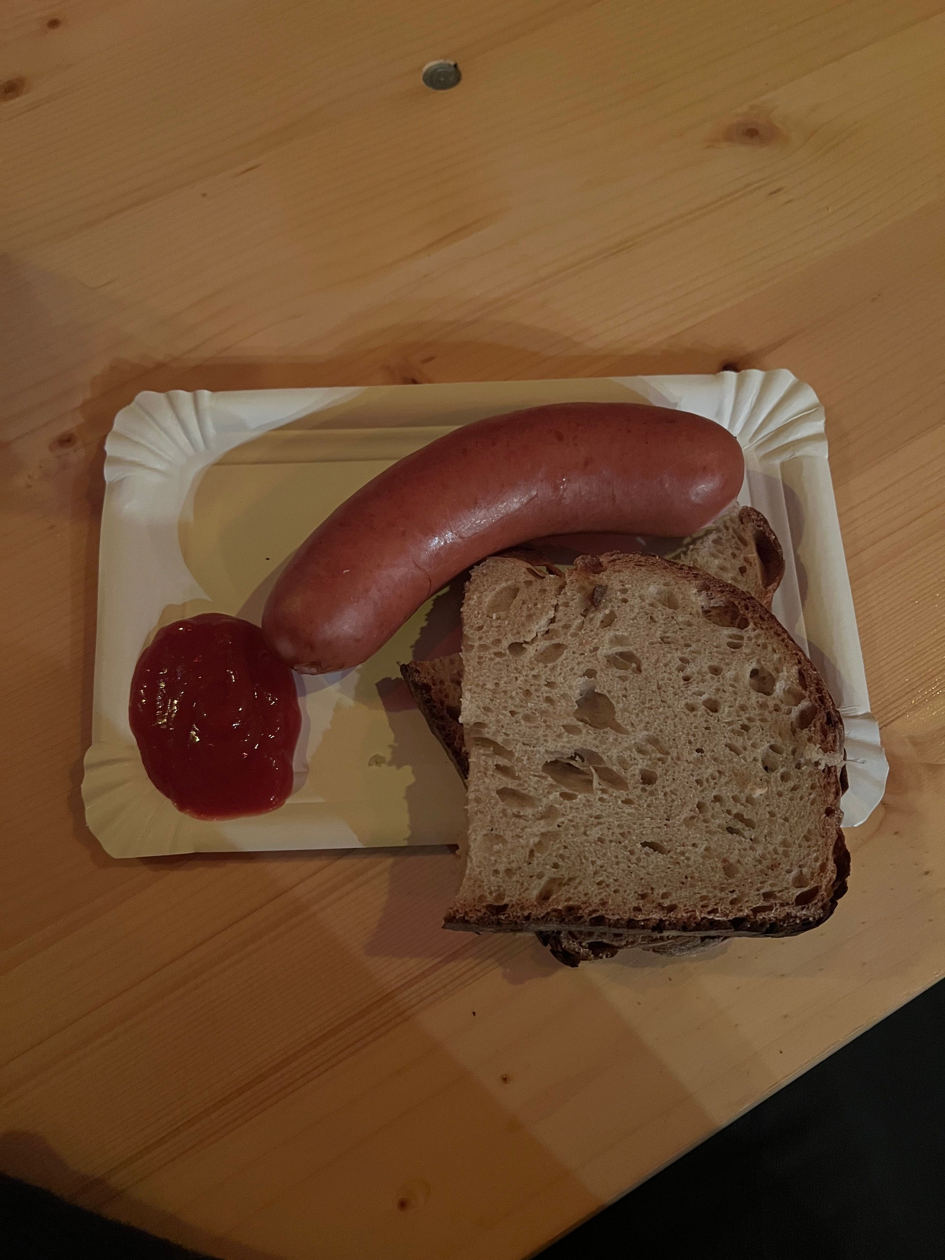 A sausage, a slice of bread, and a dollop of ketchup on a white paper plate resting on a wooden table.