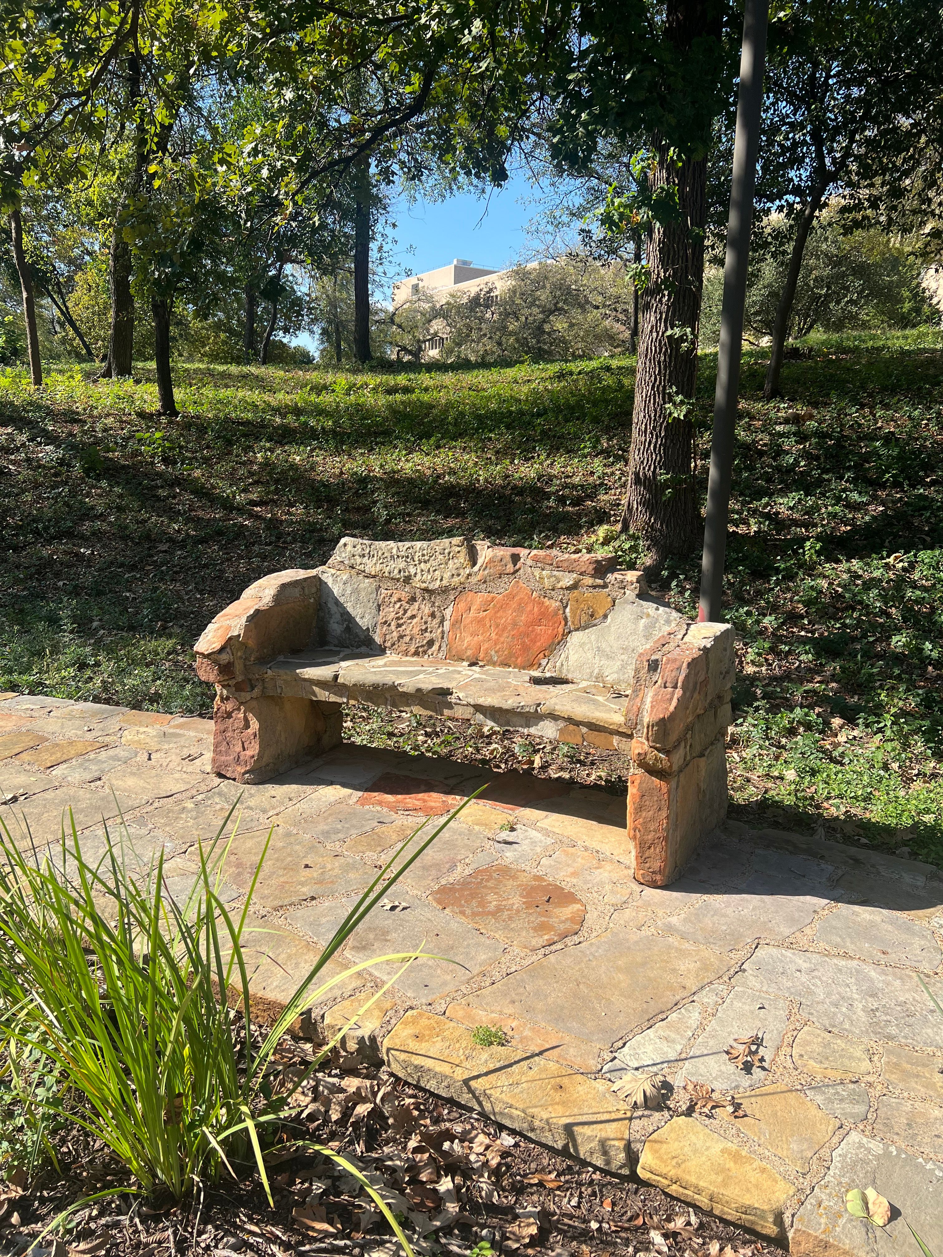 A rustic stone bench sits on a flagstone patio beneath trees on a sunlit grassy hillside, with a building visible in the background.