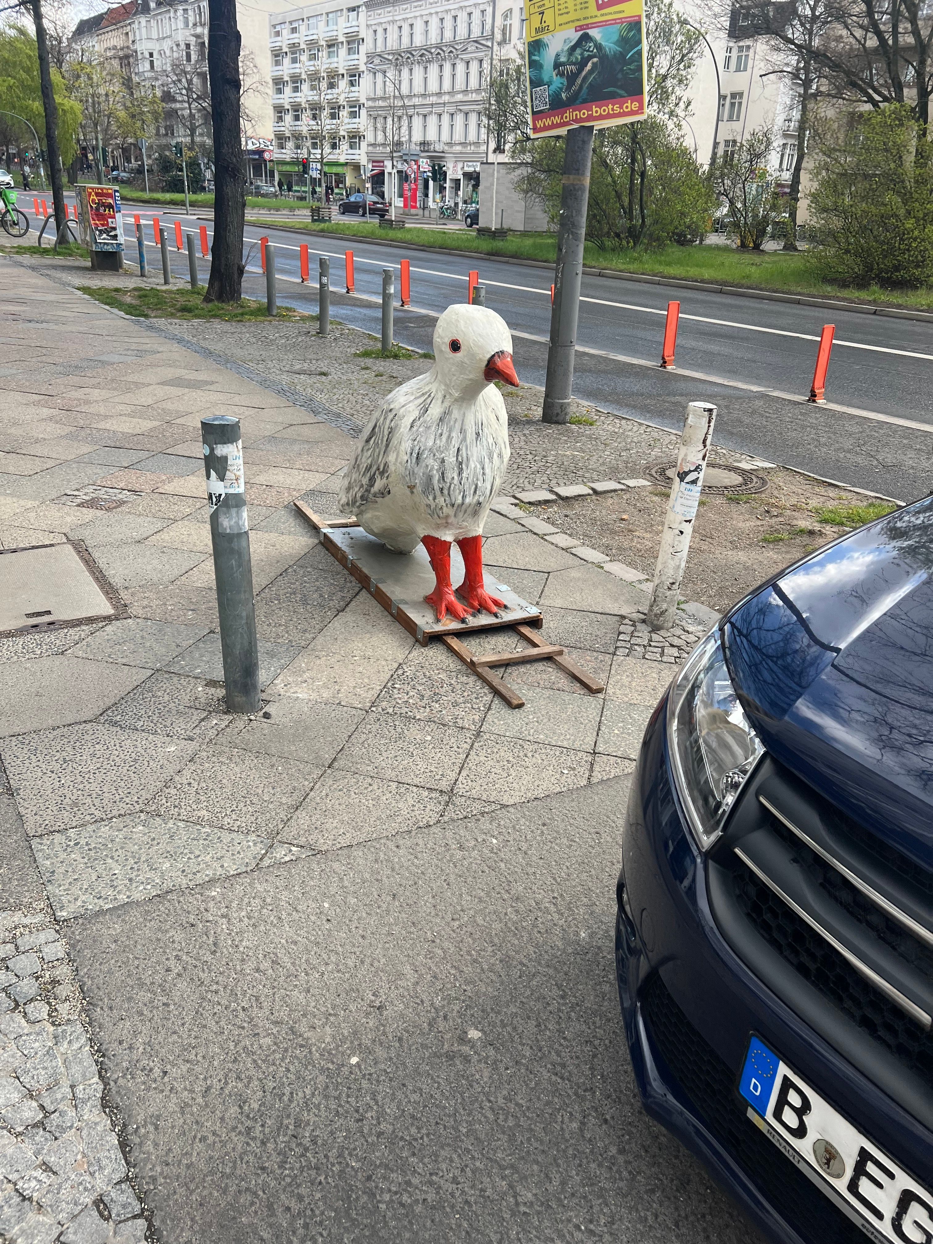 A large white bird sculpture with red beak and feet stands on a wooden pallet on a city sidewalk in Berlin, next to a metal bollard and a dark blue car, with a street and buildings in the background.