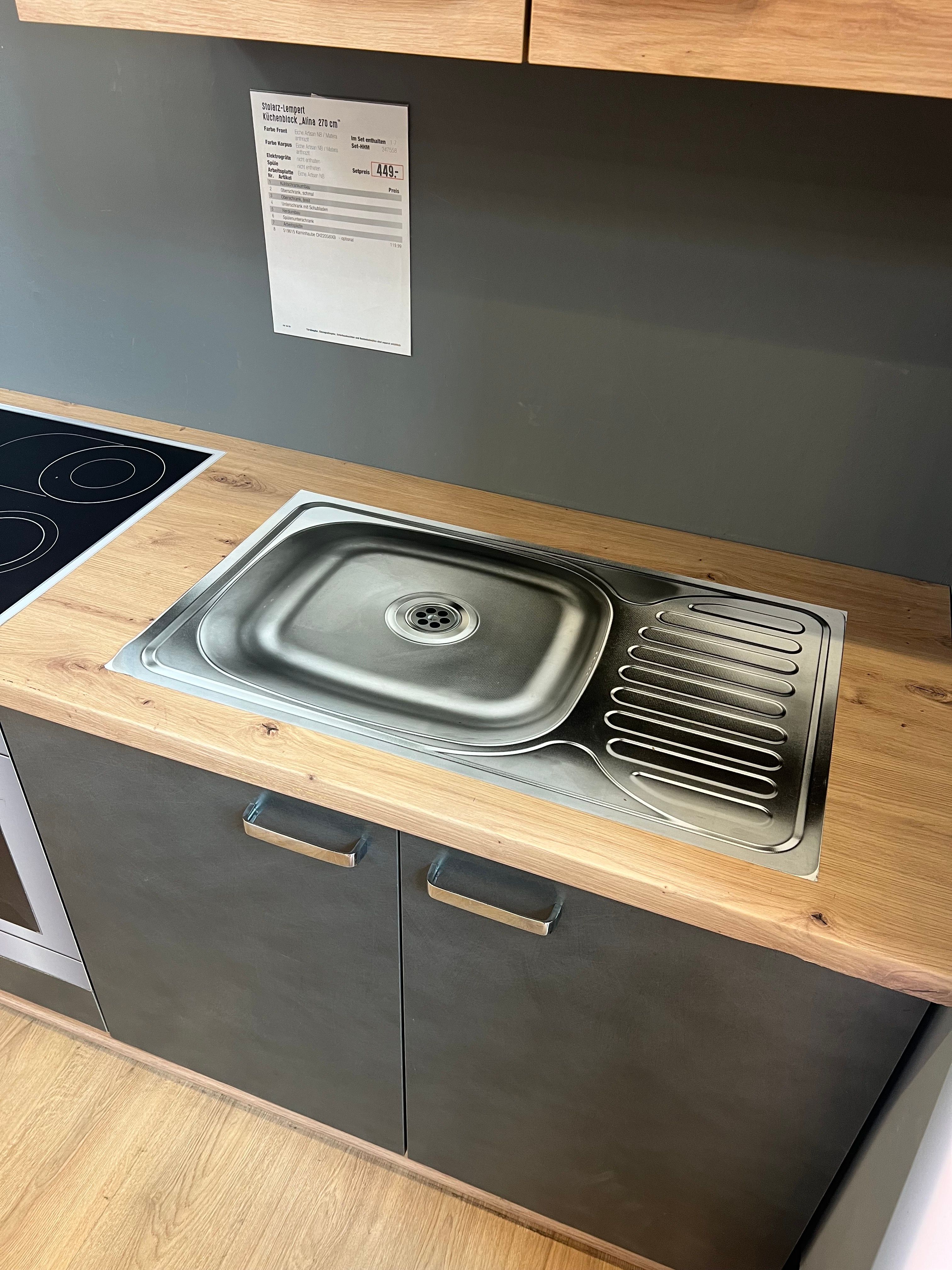 A stainless steel kitchen sink with drainboard set into a wooden countertop above gray cabinets, with a price tag on the gray wall and a black cooktop to the left.
