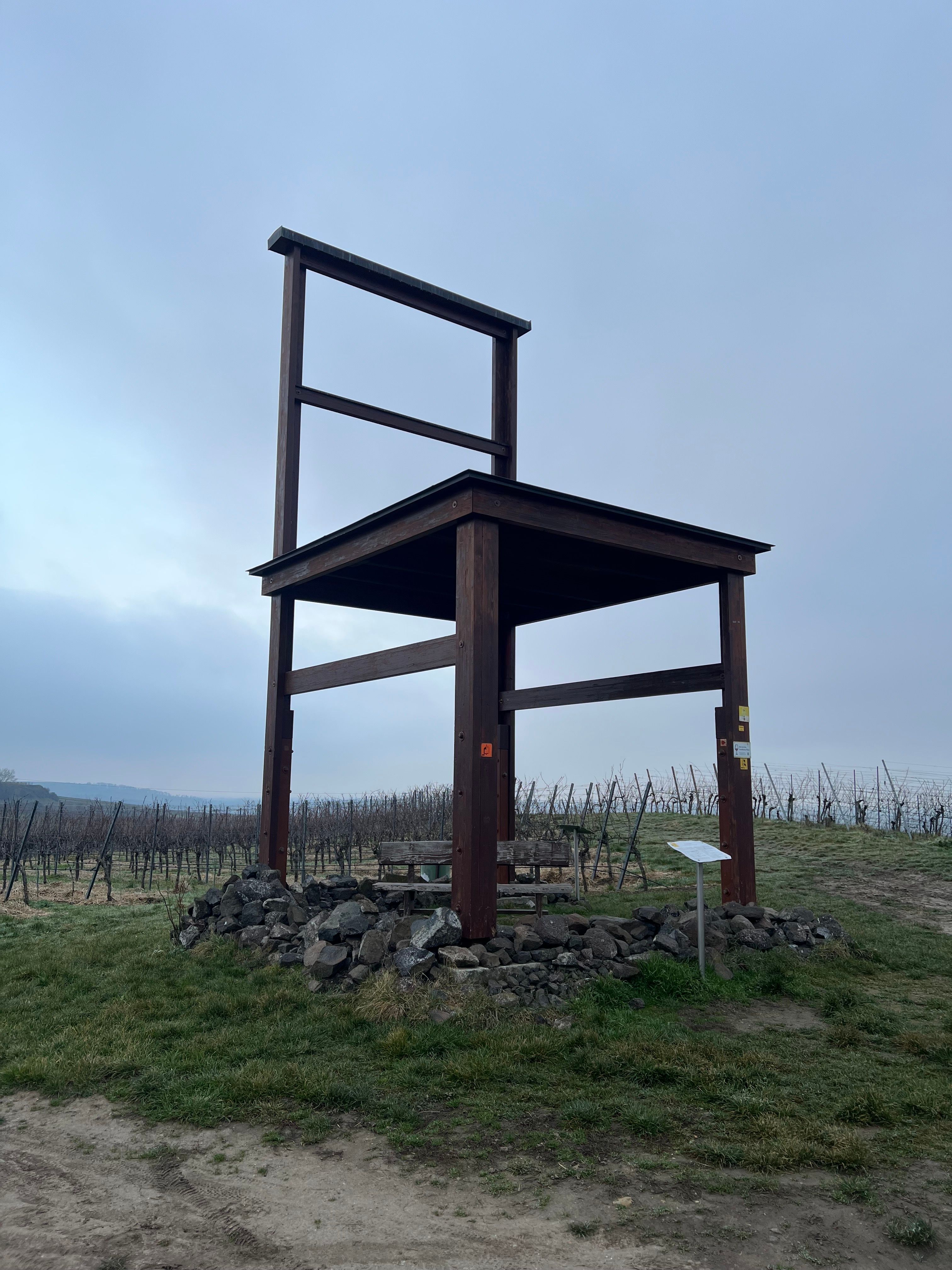 A giant wooden chair sculpture standing on a base of stones in a vineyard under an overcast sky.
