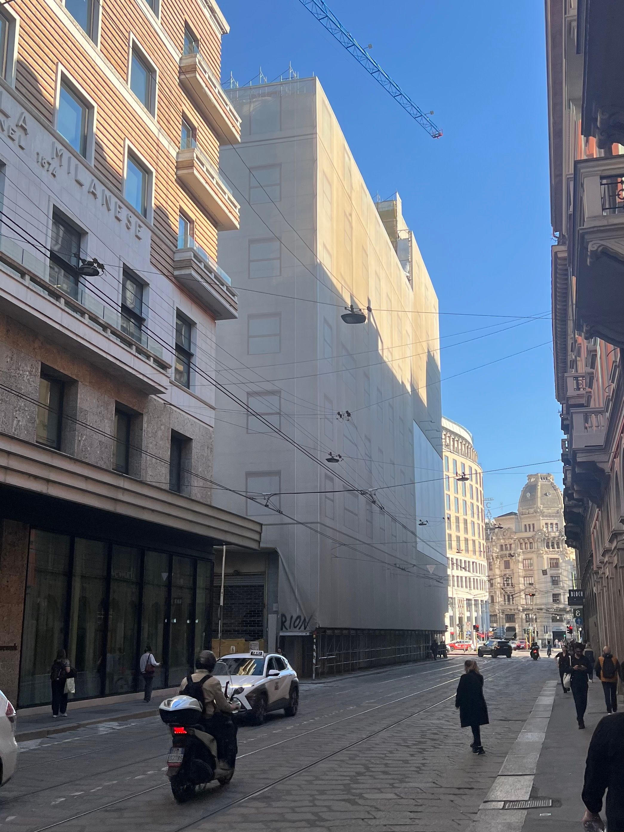 A city street in Milan with a tall building wrapped in scaffolding fabric, a construction crane overhead, and a scooter, taxi, and pedestrians on the cobblestone road.