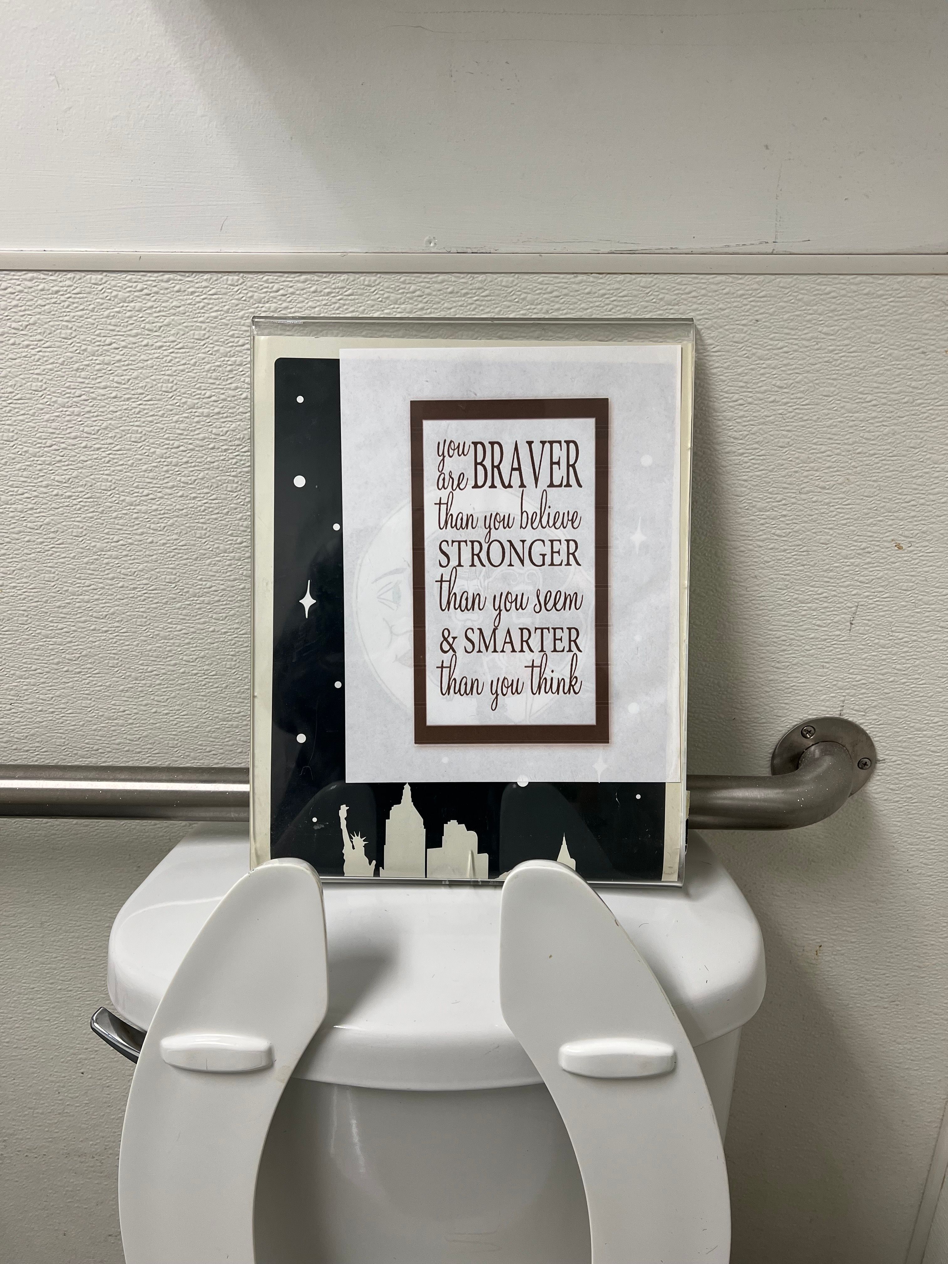 A framed motivational sign reading "you are braver than you believe, stronger than you seem & smarter than you think" propped on top of a white toilet next to a metal grab bar in a restroom.