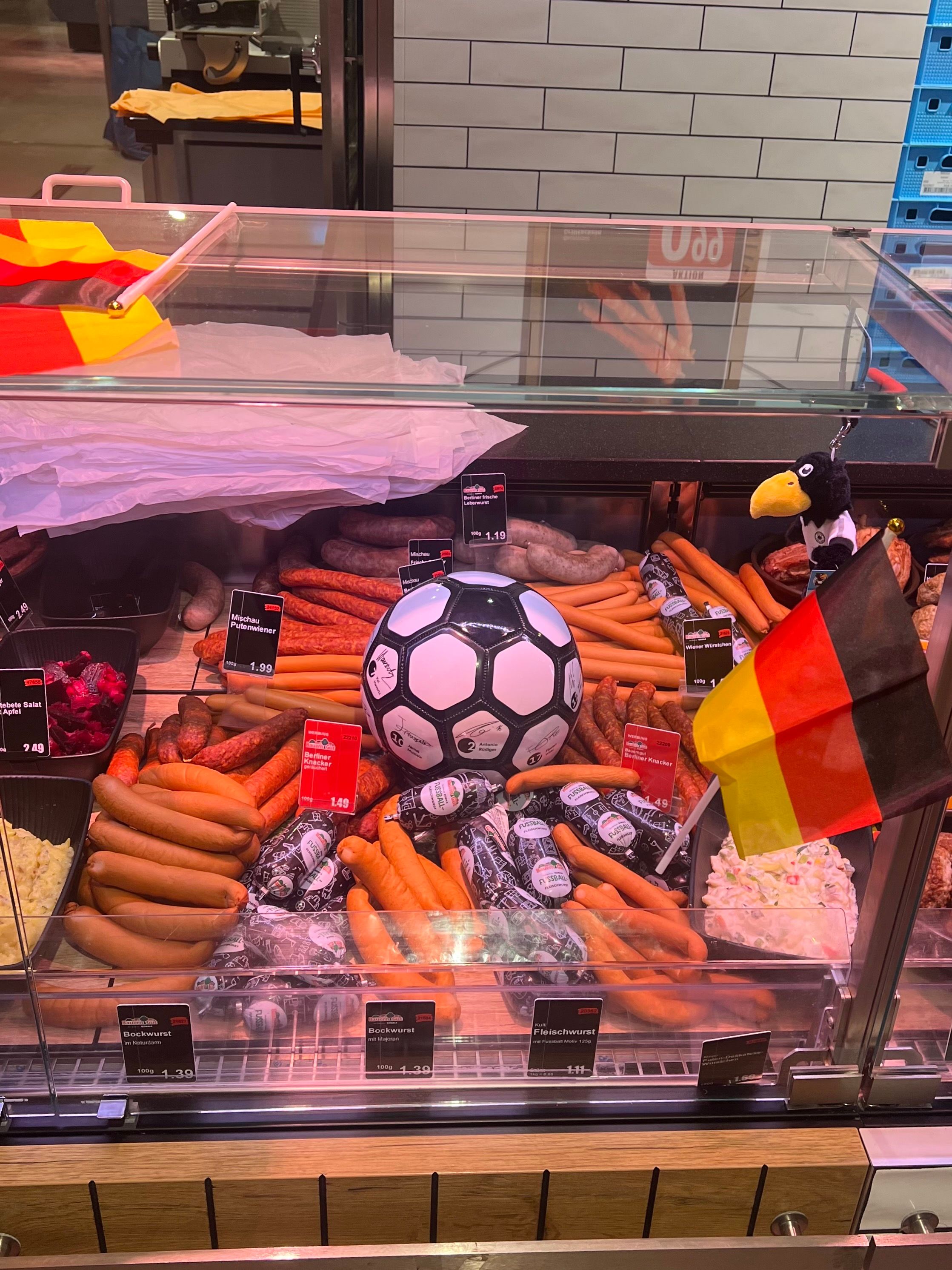 A butcher's display counter filled with various sausages arranged around a soccer ball, with a German flag and a plush eagle mascot beside it.