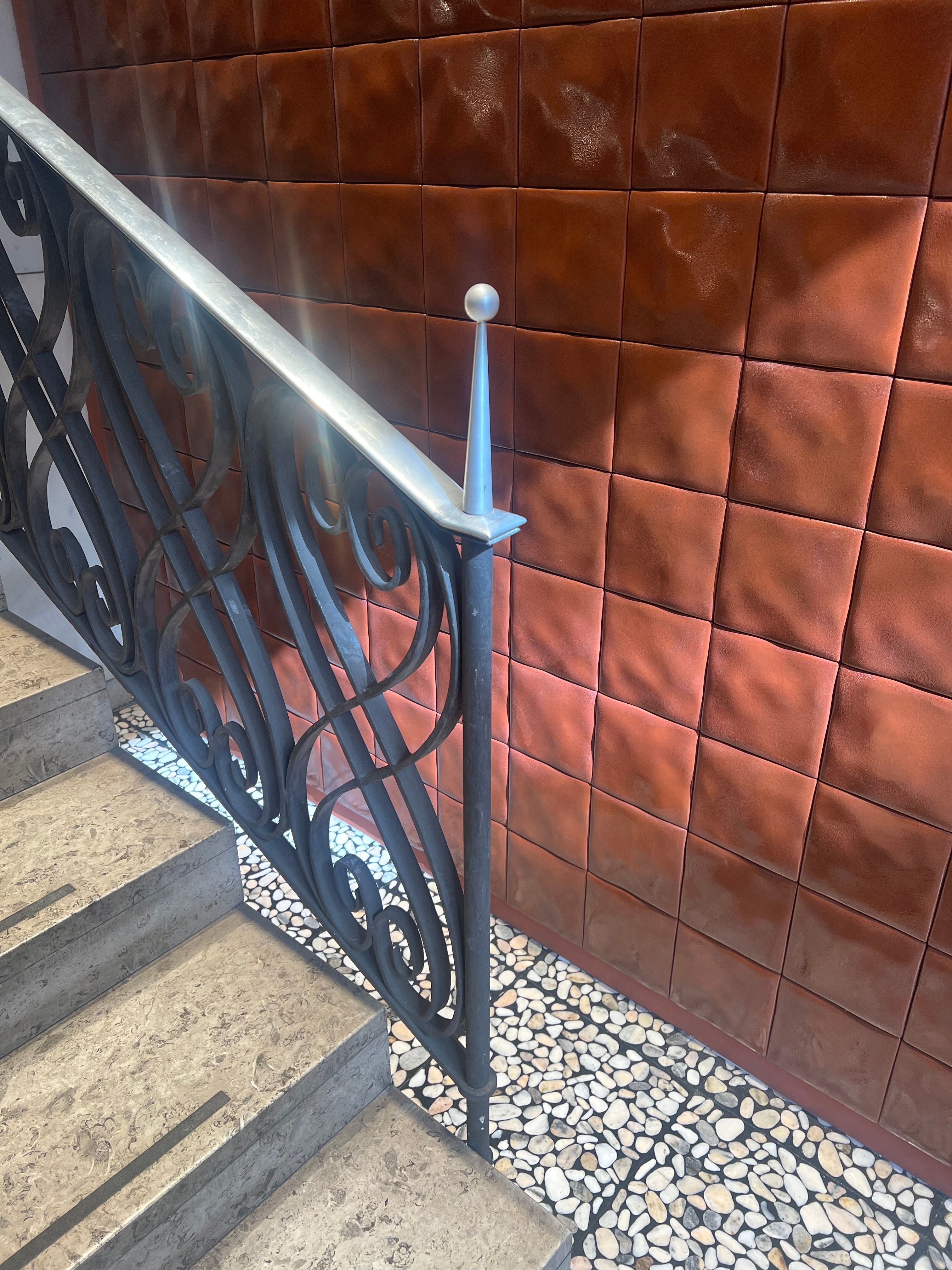 An ornate black wrought-iron stair railing with a metal handrail and pointed finial stands beside a glossy reddish-brown tiled wall above a pebbled terrazzo floor.
