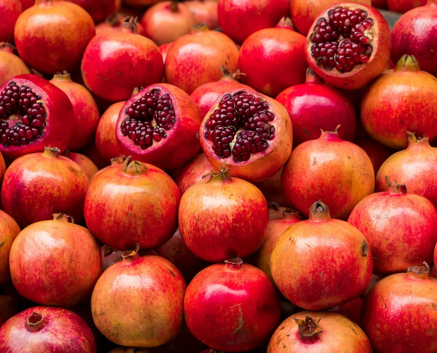 A large pile of fresh pomegranates, some of which are cut open to reveal their vibrant, red, juicy seeds.