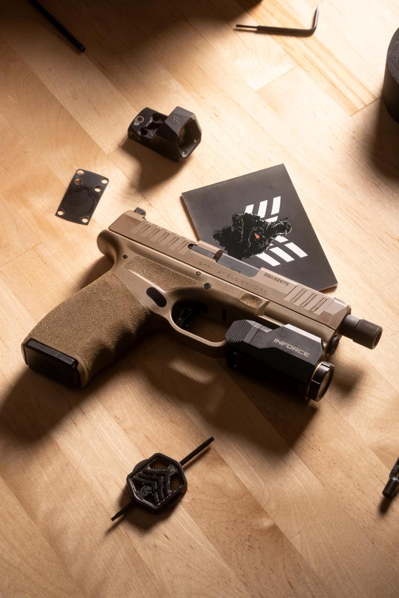 FDE Springfield Hellcat Pro with threaded barrel and weapon light on a workbench beside a Swampfox Sentinel II and mounting plate