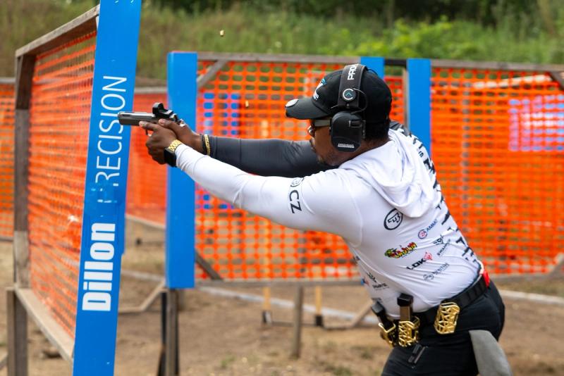 DJ Jenkins in full stride, pistol extended, sponsor logos visible on his shirt, next to a Dillon Precision sign