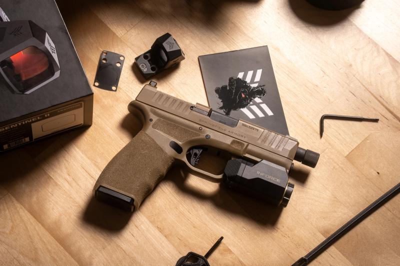 FDE Springfield Hellcat Pro with weapon light next to a Swampfox Sentinel II red dot, its box, and a mounting plate on a workbench