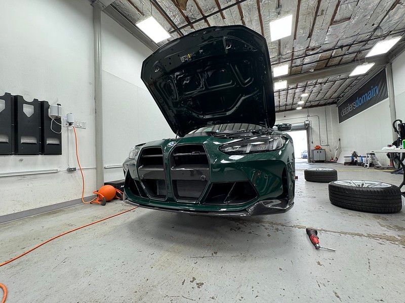 Front view of a dark green BMW G80 M3 with hood raised and wheels removed inside the Detailer's Domain shop, being prepped for paint protection film installation.