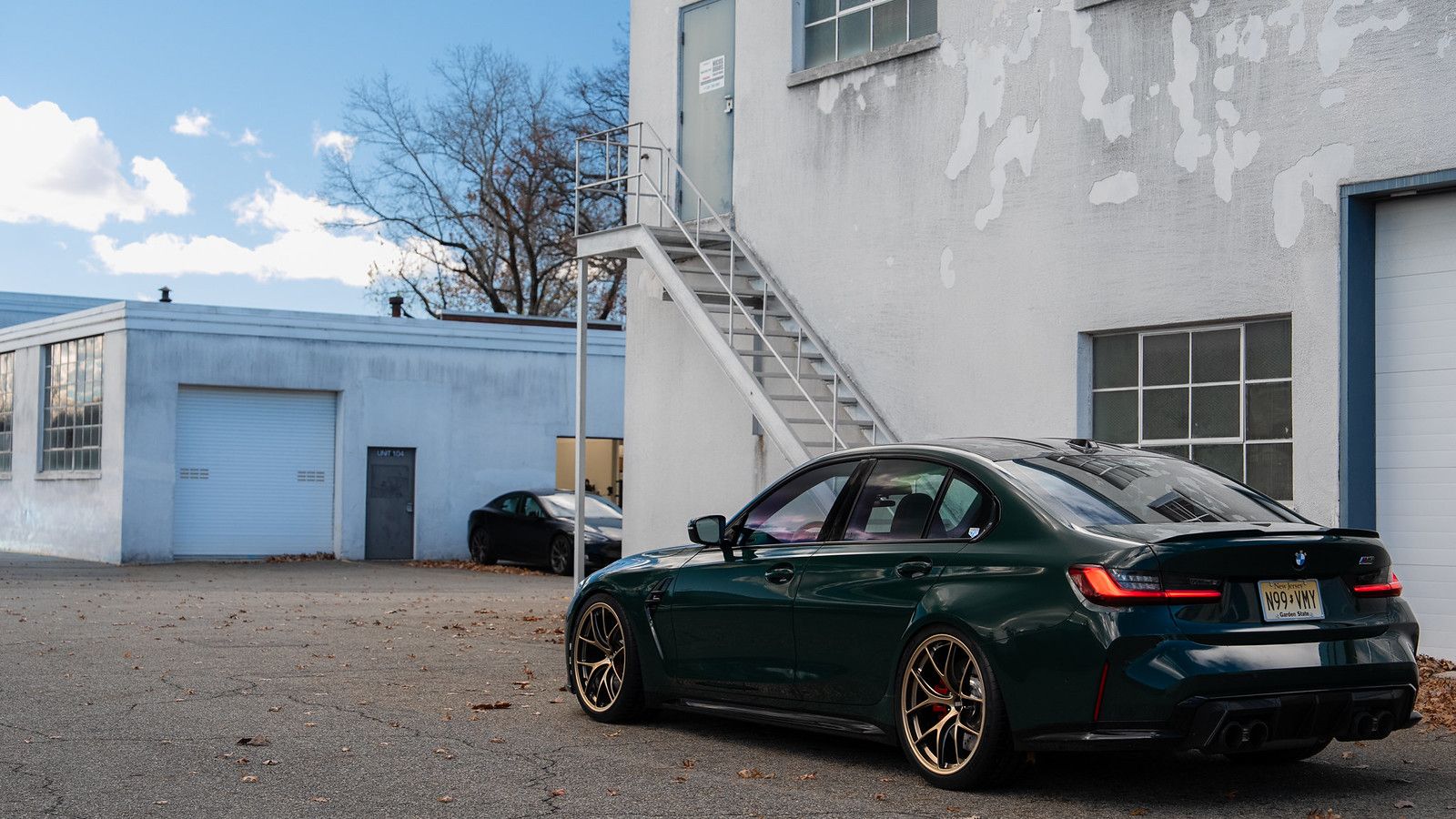 Rear three-quarter view of a lowered British Racing Green BMW G80 M3 with bronze wheels parked outside the shop.