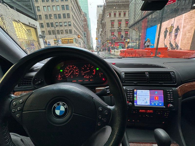 View from the driver's seat of a BMW E38 7 Series showing a freshly detailed interior with wood trim, illuminated gauges, and an aftermarket Apple CarPlay head unit while driving through a city.