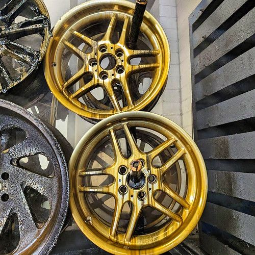 Two BMW M-Parallel style wheels freshly refinished in a gold powder coat finish, displayed alongside unpainted wheels in a workshop.