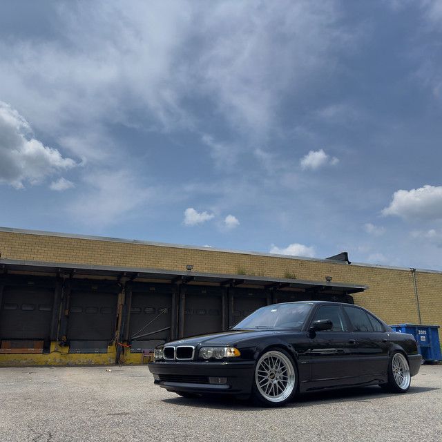 Lowered black BMW E38 7 Series with aftermarket mesh wheels parked in front of an industrial warehouse under a dramatic cloudy sky.