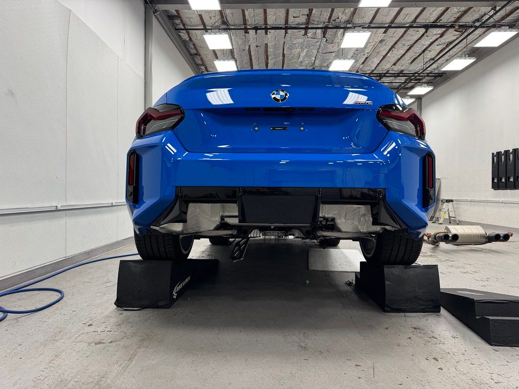 Rear view of a Zandvoort Blue BMW M2 raised on ramps in a brightly lit detail shop with bumper and trunk removed for PPF installation.