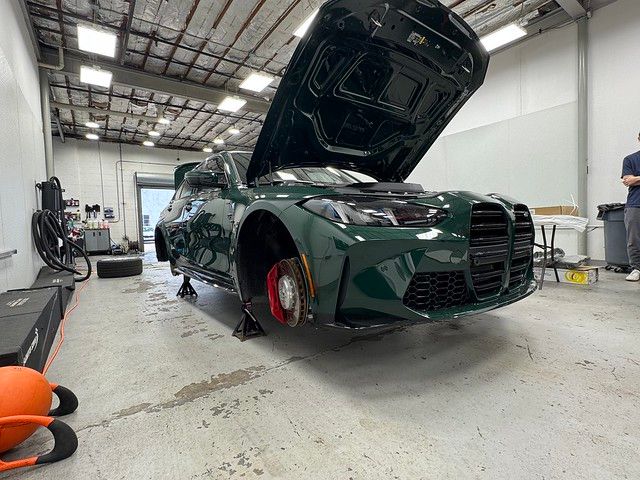 British Racing Green BMW G80 M3 on jack stands with wheels removed and hood open inside the detailing shop during a full build.