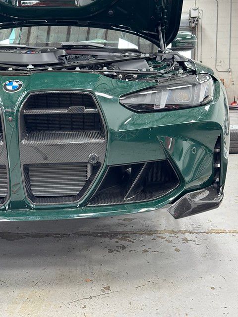 Close-up of a British Racing Green BMW M3 front end with carbon fiber front lip, canards, and intake surrounds under shop lighting.