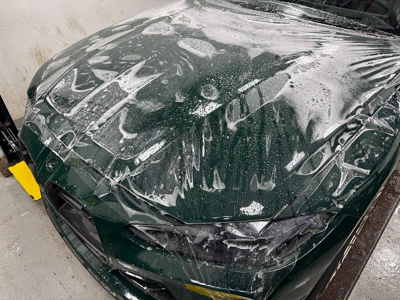 Close-up of a dark green BMW M3 hood during PPF installation showing clear film with soapy slip solution being worked across the painted surface.