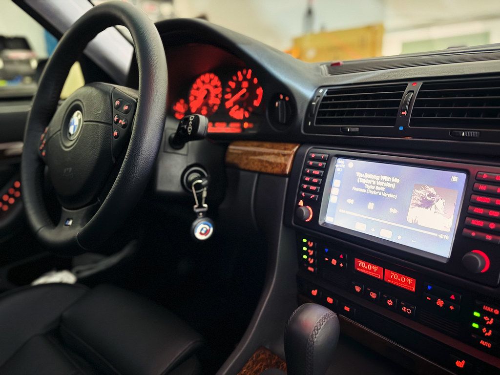 Interior view of a BMW E38 showing the steering wheel, red-lit instrument cluster, wood trim, and aftermarket touchscreen head unit.