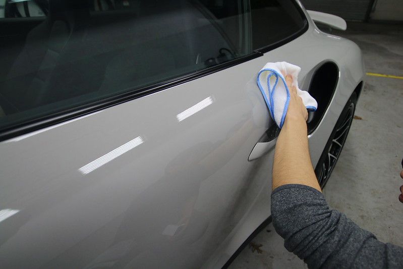 A detailer hand-wiping the side panel of a silver Porsche 911 with a microfiber cloth, revealing a deep glossy finish with reflected shop lights.