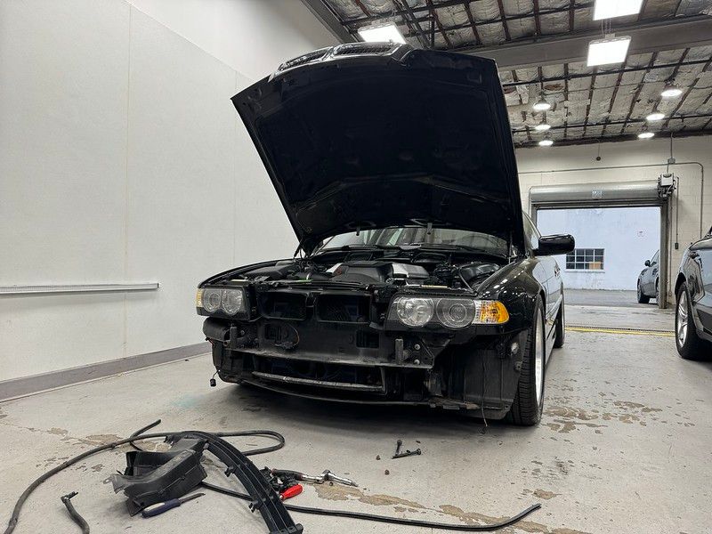 Black BMW E38 7 Series with hood open and front bumper removed in the shop, being prepped for paint protection film or paint correction work.