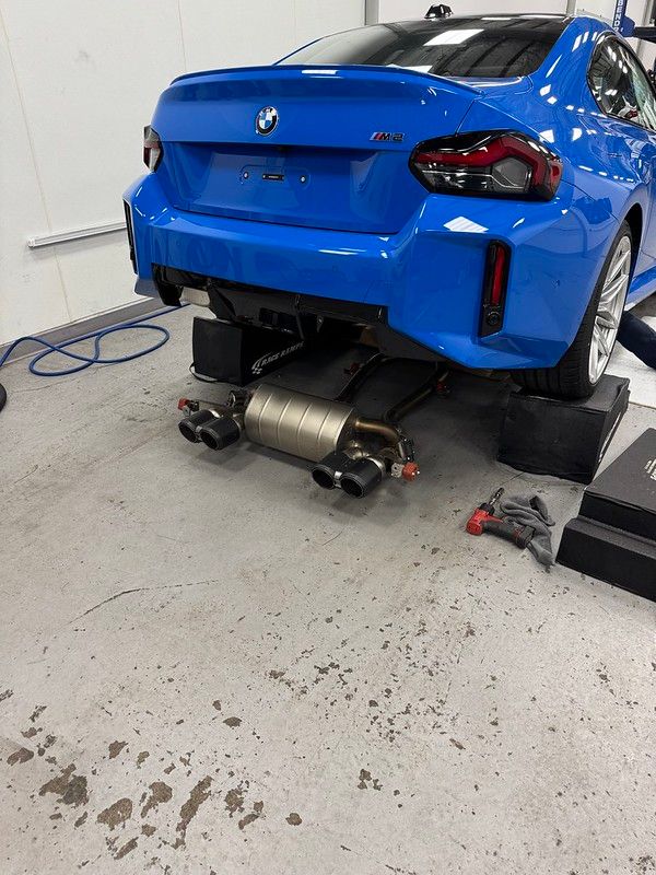 Rear view of a Zandvoort Blue BMW G87 M2 on jack stands in the shop with an aftermarket exhaust system being installed underneath.