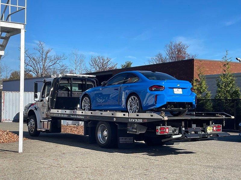 Zandvoort Blue BMW G87 M2 on a flatbed truck being delivered to the shop for paint protection film installation on a sunny day.