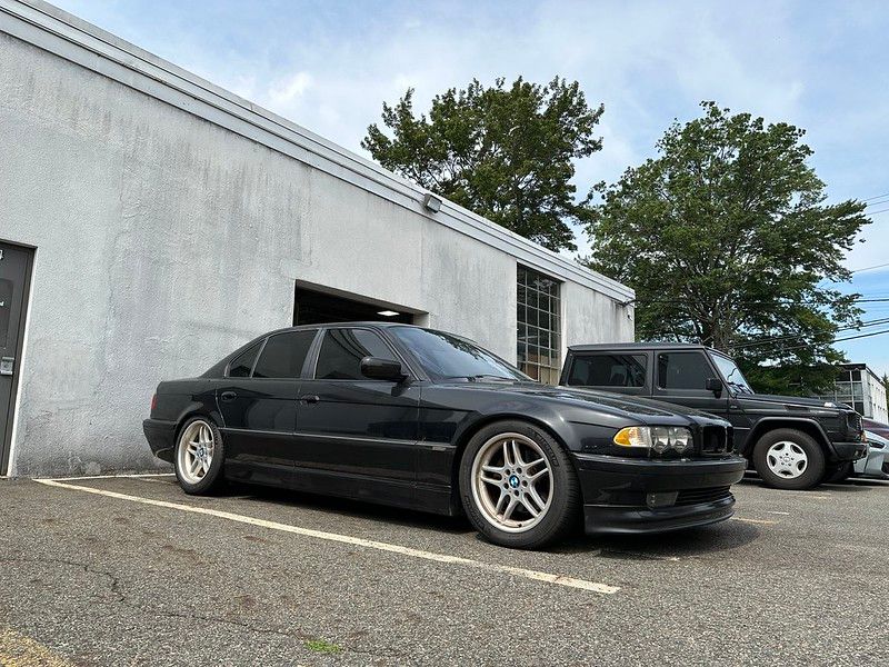 Black BMW E38 7 Series with glossy paint and polished M-Parallel wheels parked outside the Detailer's Domain shop after a full detail, with a Mercedes G-Wagon visible alongside.
