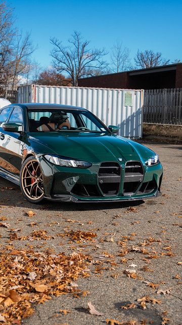 Front three-quarter view of a lowered Isle of Man Green BMW G80 M3 with gold wheels and carbon accents, parked on pavement with autumn leaves.