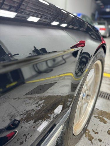 Extreme close-up of a black BMW E38 rear quarter panel reflecting shop lights, demonstrating a flawless mirror-like paint correction finish.