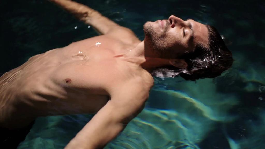A man floating on his back in a pool, eyes closed, in rippling light.