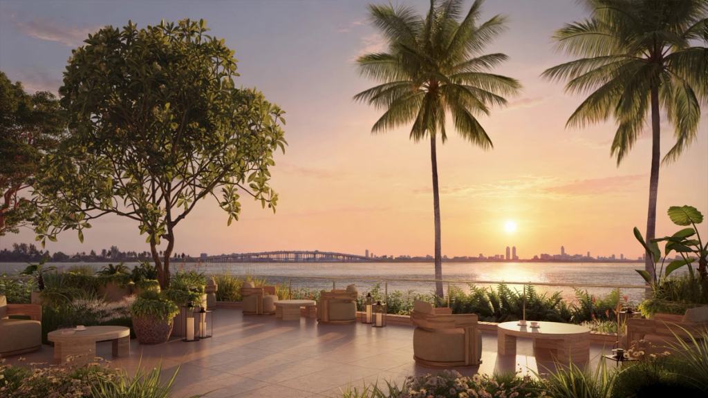 A landscaped waterfront terrace with lounge seating and tropical greenery overlooking calm water and a city skyline at sunset
