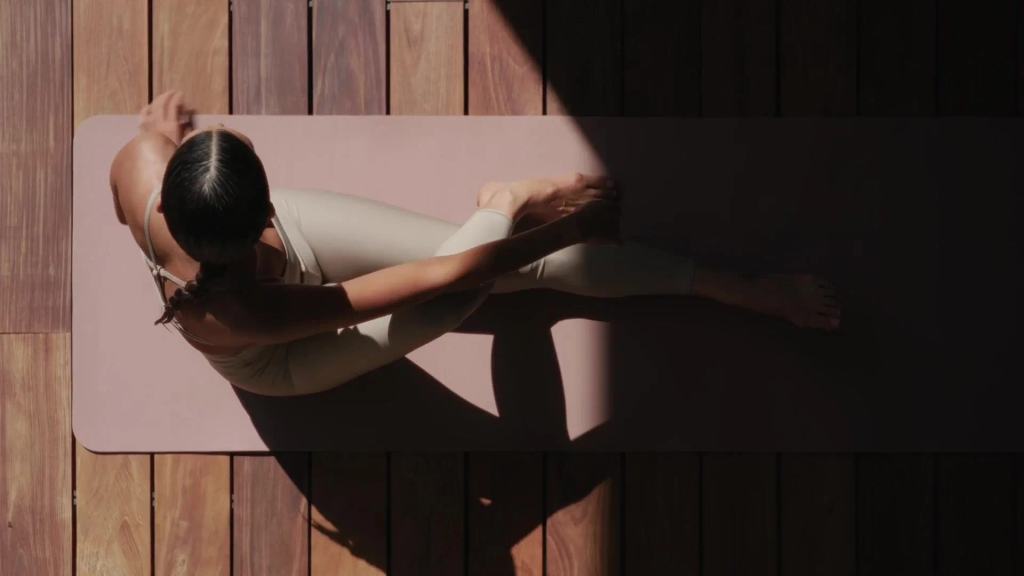 Overhead view of a woman stretching on a yoga mat on a sunlit wood deck.