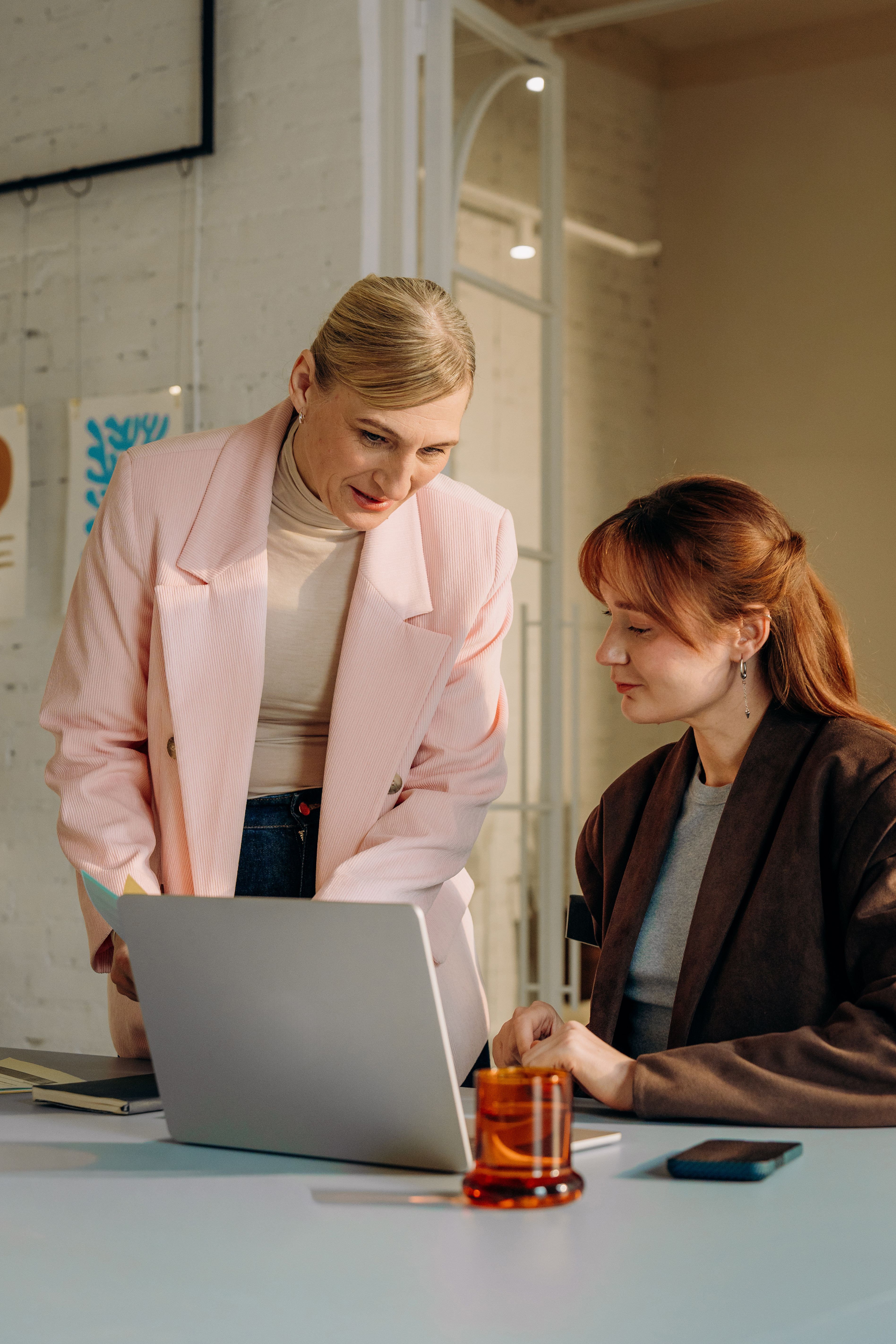 Two women, one standing and one seated, looking at a laptop together.
