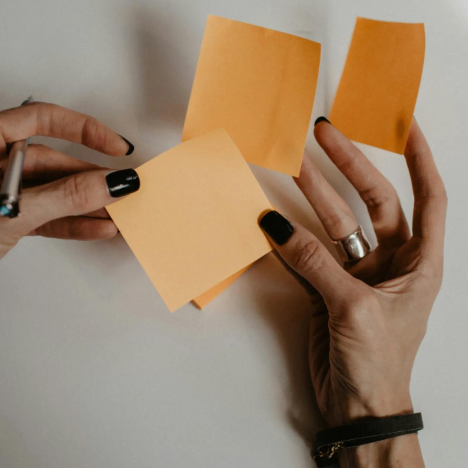 Hands hold several blank orange sticky notes over a neutral surface