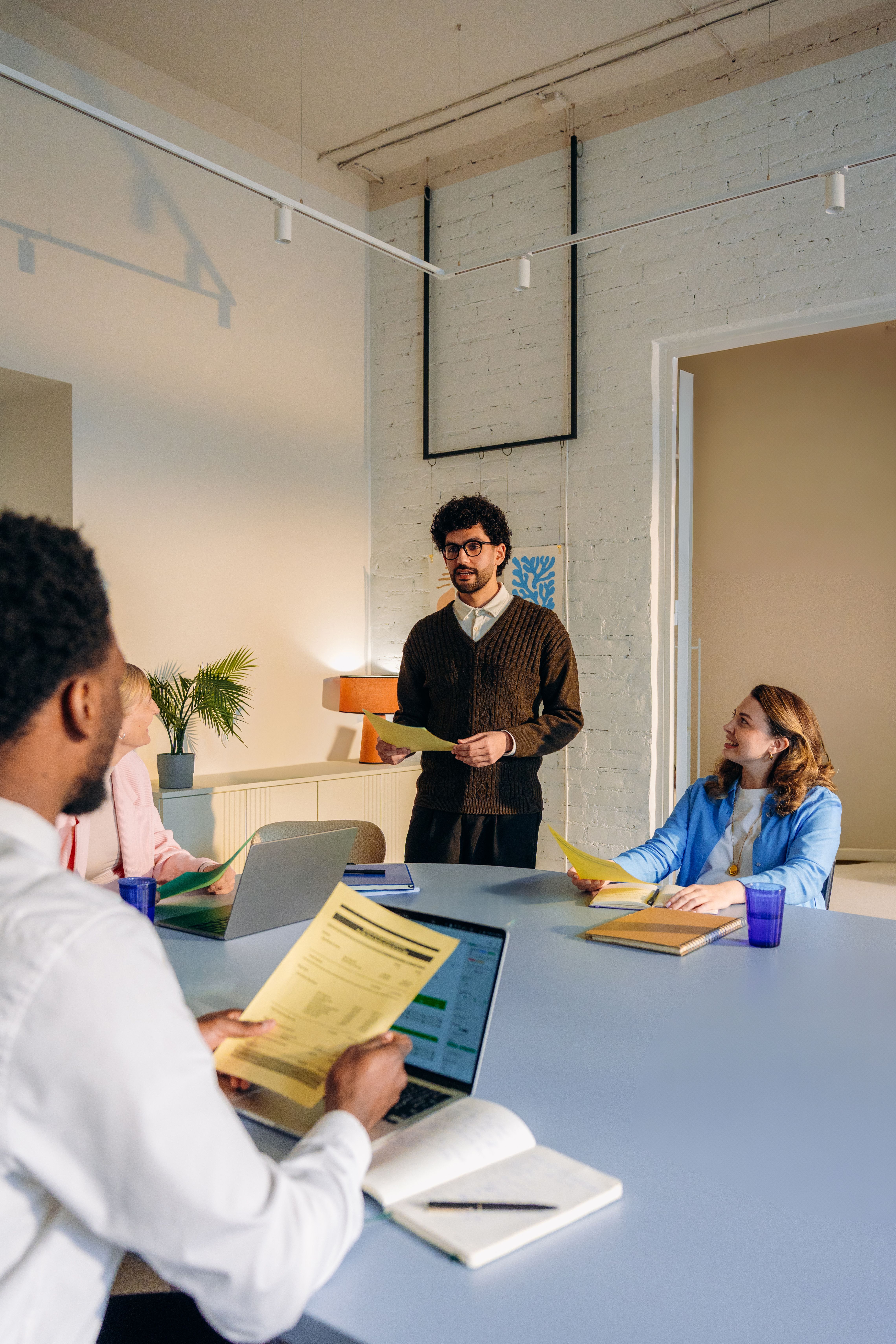 A man presents yellow papers to three colleagues seated at a conference table with laptops.