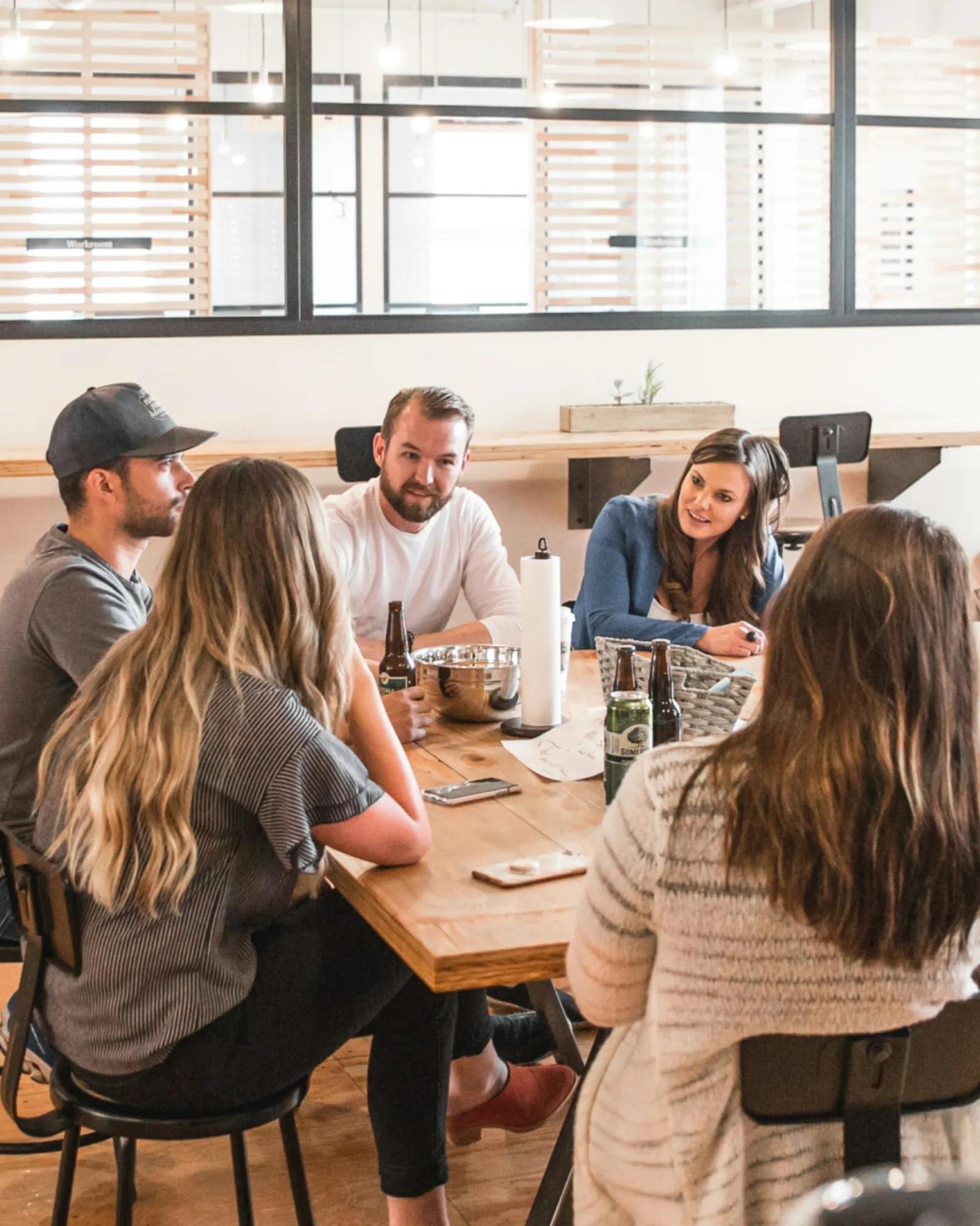 A small group of people sit around a wooden table in a modern office, talking and collaborating during an informal meeting, with notebooks, drinks, and laptops on the table.